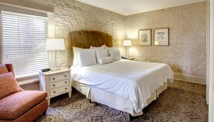 This is a well-lit cozy bedroom featuring a large bed with white linen exposed white brick walls elegant furniture and framed artwork