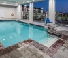 The image shows a calm and inviting indoor swimming pool area during the evening with clear blue water a handicap-accessible lift chair and an outdoor view through large windows