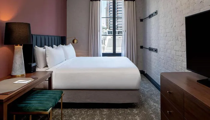 The image shows a modern bedroom with a neatly made bed a white painted brick wall and a city view through the window