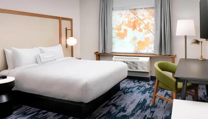 The image shows a neatly arranged hotel room with a large bed a desk with a chair and a window showcasing a view of autumn leaves