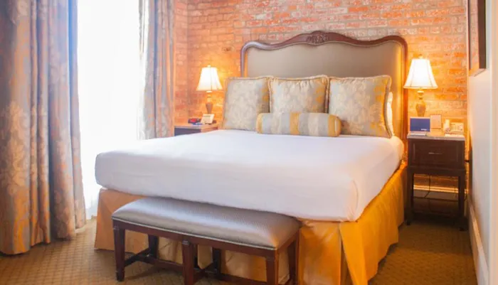 The image shows a cozy hotel room with a large bed against an exposed brick wall accented with elegant bedding and classic furniture