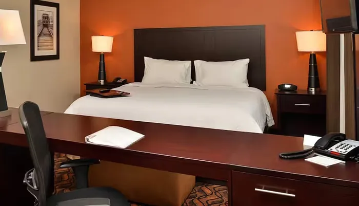 The image shows a neatly arranged hotel room with a large bed a desk with a chair and various items like a phone lamps and decorative elements