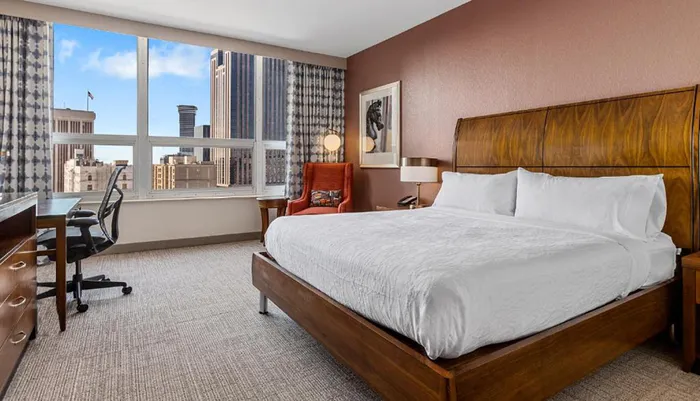 The image shows a neatly arranged modern hotel room with a large window offering a city view a comfortable bed work desk and elegant interior decor