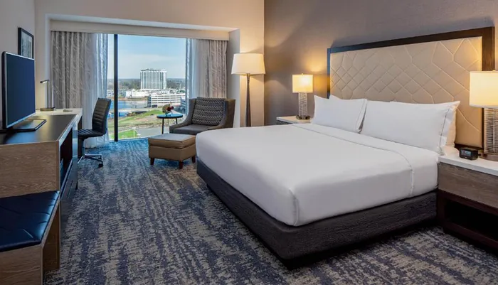 The image shows a neatly arranged hotel room with modern furniture and a view of a cityscape outside the window