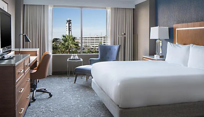 The image shows a modern and neatly arranged hotel room with a large bed a desk seating areas and a window offering a view of a city landscape and a distinctive tower