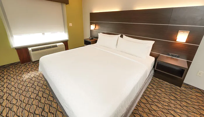 The image shows a neatly made bed with white bedding in a modern hotel room with a patterned carpet simple furnishings and warm lighting