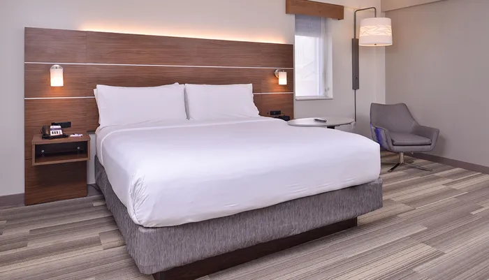 The image shows a neatly arranged modern hotel room with a large bed an armchair side tables and tasteful lighting