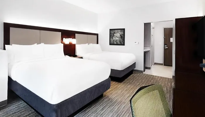 This image shows a modern and neatly arranged hotel room with two queen-sized beds clean white linens a wall-mounted art piece and a glimpse of the entrance area