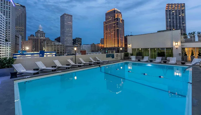 The image shows a tranquil rooftop swimming pool flanked by lounge chairs with a backdrop of the city skyline during dusk