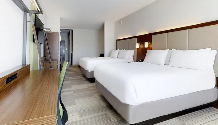 The image shows a modern hotel room with two large beds crisp white bedding minimalist furniture and a bright clean interior