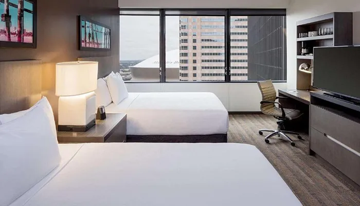 This image shows a modern hotel room with two beds a work desk a large window with a city view and contemporary furnishings