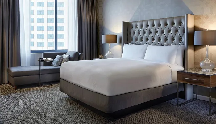 This image shows a neatly arranged modern hotel room with a large bed elegant furniture and a window with a view of a city building