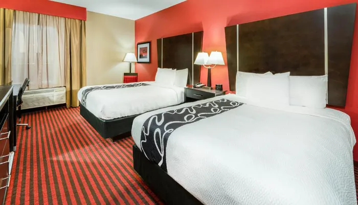 The image shows a neatly arranged hotel room with two queen-sized beds featuring a red and black color scheme and a striped carpet