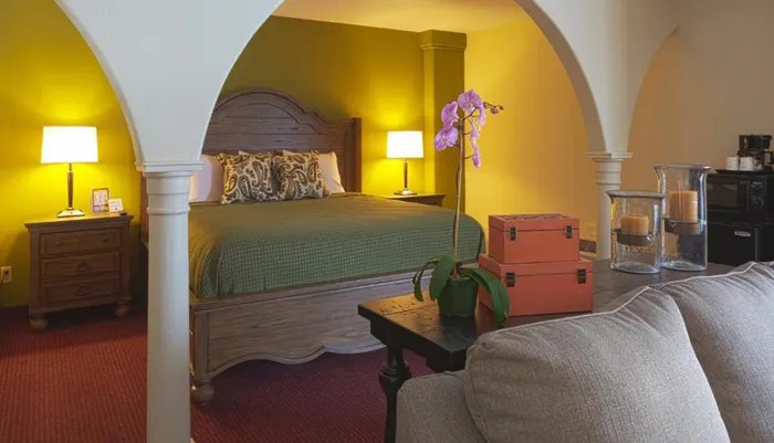 The image shows a cozy and warmly lit bedroom with a green bedspread wood furniture a bright yellow accent wall and decorative elements like an orchid and orange storage boxes