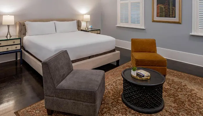 An elegantly decorated bedroom features a large bed with white linens flanked by nightstands with lamps complemented by a gray armchair a mustard accent chair and a round black coffee table set on a patterned area rug