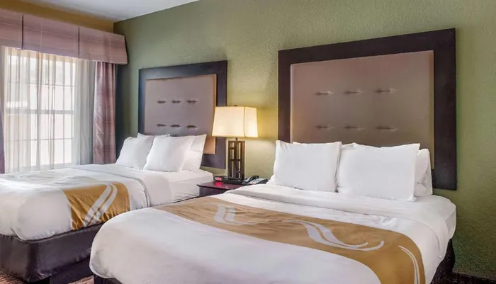 The image shows a well-lit hotel room with two neatly made beds featuring white linens and decorative gold runners flanked by nightstands and an upholstered headboard against a green wall