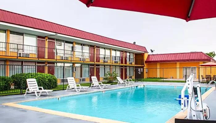 This image shows a two-story motel with exterior corridors overlooking a small outdoor swimming pool and lounge chairs with a bold color scheme and a clear sky