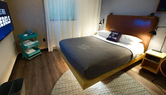 The image shows a modern and stylishly decorated hotel room featuring a neatly made bed with a decorative pillow a small trolley with towels a flat-screen TV and warm lighting