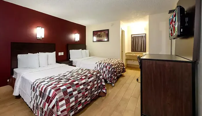 The image shows a modestly furnished hotel room with two beds covered in plaid-patterned bedspreads a mounted television and a simple decor with a burgundy accent wall