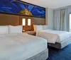 A modern hotel room features two neatly made double beds an artistic wall panel above table lamps between beds and a city view through the large window