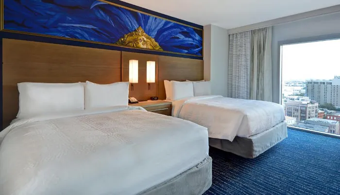 A modern hotel room features two neatly made double beds an artistic wall panel above table lamps between beds and a city view through the large window