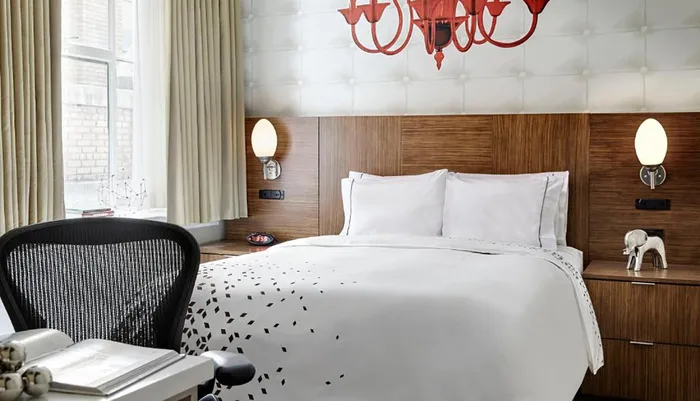 The image presents a modern and stylish hotel room with an artistic red chandelier a patterned bedspread wooden headboard and tasteful decor
