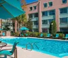 The image shows a sunny poolside area with loungers umbrellas and a clear blue swimming pool at what appears to be a hotel or resort
