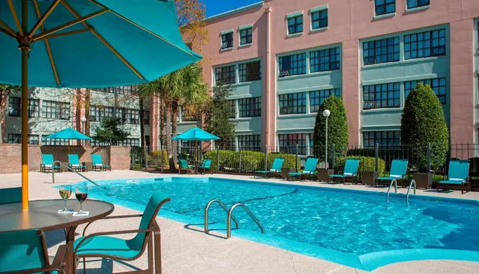 The image shows a sunny poolside area with loungers umbrellas and a clear blue swimming pool at what appears to be a hotel or resort