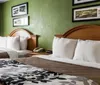 The image shows a tidy hotel room with two beds adorned with patterned bedding green walls framed artwork and sheer curtains over a window
