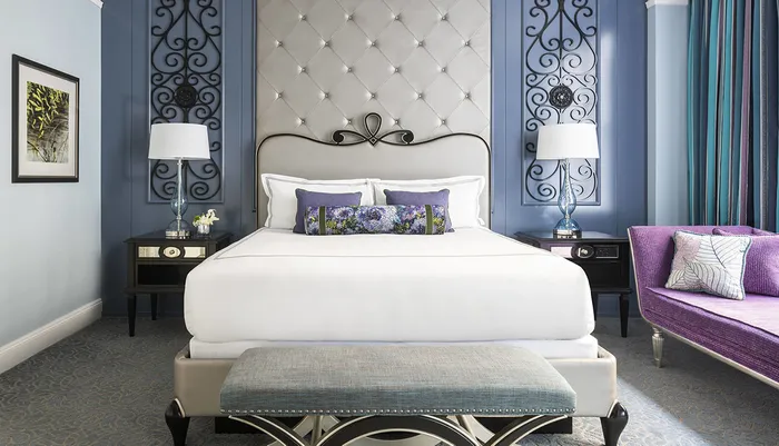 The image shows an elegantly designed bedroom featuring a plush bed with decorative pillows sophisticated furniture and a color scheme dominated by shades of blue and purple