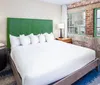 The image shows a modern bedroom with a large bed featuring a bright green headboard exposed brick wall and a city view from the window