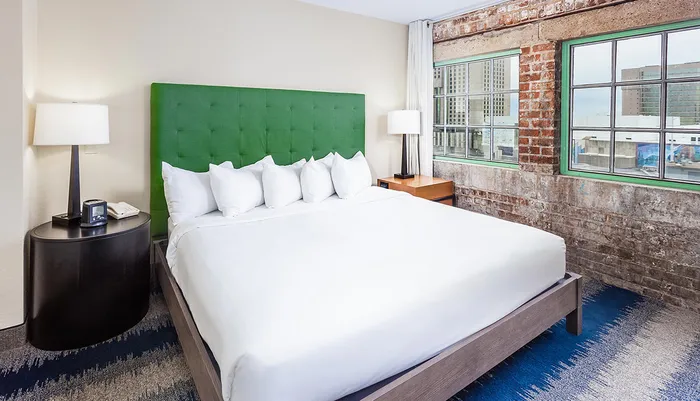 The image shows a modern bedroom with a large bed featuring a bright green headboard exposed brick wall and a city view from the window