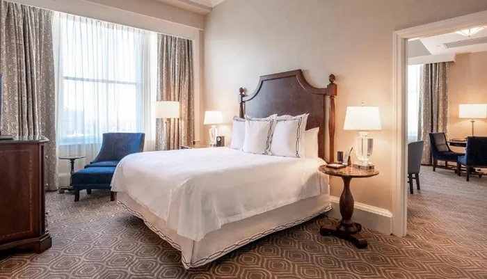 The image shows an elegantly furnished hotel room with a large bed ornate headboard sitting area and a patterned carpet