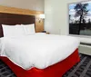 This is a modern hotel room with a large bed featuring white bedding and a red bed skirt a wooden headboard with attached lights a dresser with a phone and a window offering a view of trees and a body of water