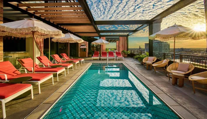 The image shows a rooftop pool area with red loungers and fringed umbrellas during sunset offering a picturesque city view