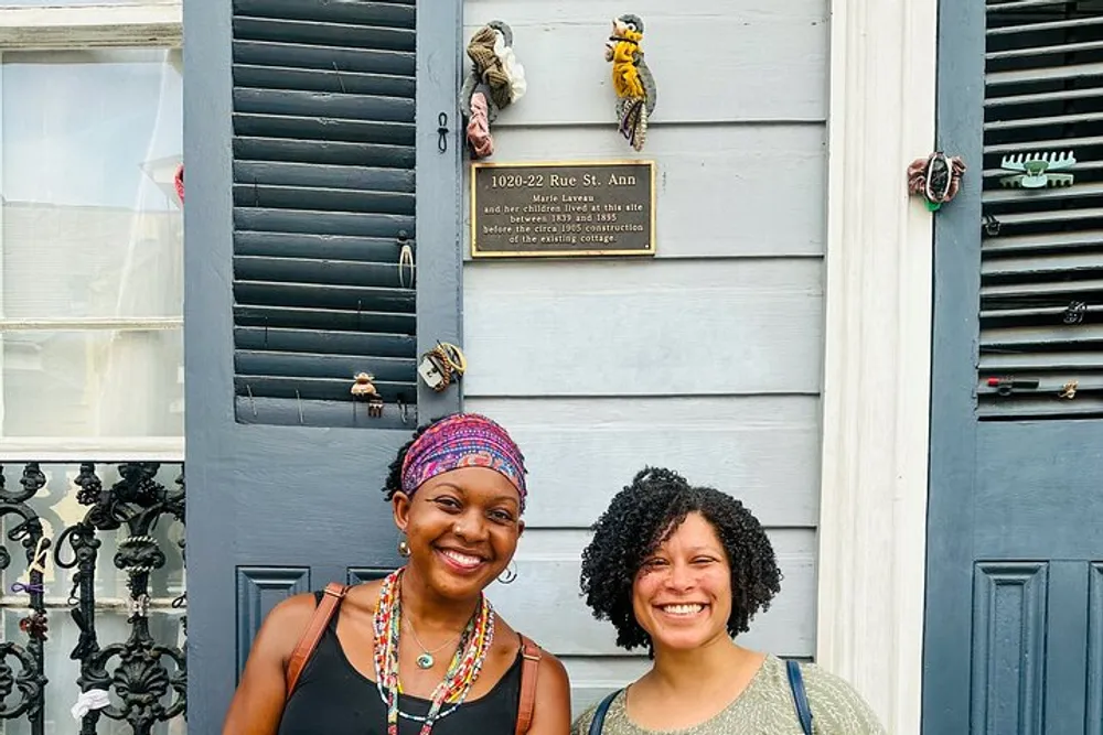 Two smiling individuals stand in front of a gray door that displays a historical plaque and decorative door knockers