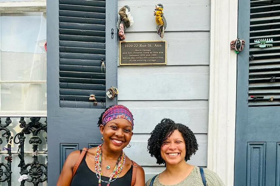 Two smiling individuals stand in front of a gray door that displays a historical plaque and decorative door knockers.