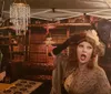 A person dressed in vintage attire is posing dramatically in front of a backdrop with a chandelier and bookshelf print at what appears to be an evening event under a tent with string lights