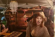 A person dressed in vintage attire is posing dramatically in front of a backdrop with a chandelier and bookshelf print, at what appears to be an evening event under a tent with string lights.