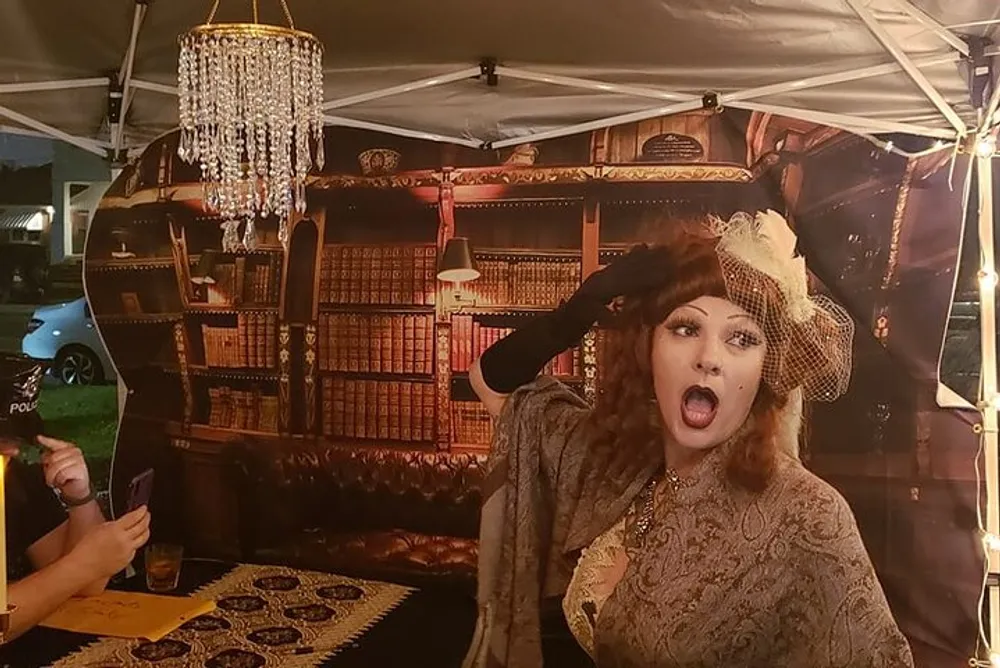 A person dressed in vintage attire is posing dramatically in front of a backdrop with a chandelier and bookshelf print at what appears to be an evening event under a tent with string lights
