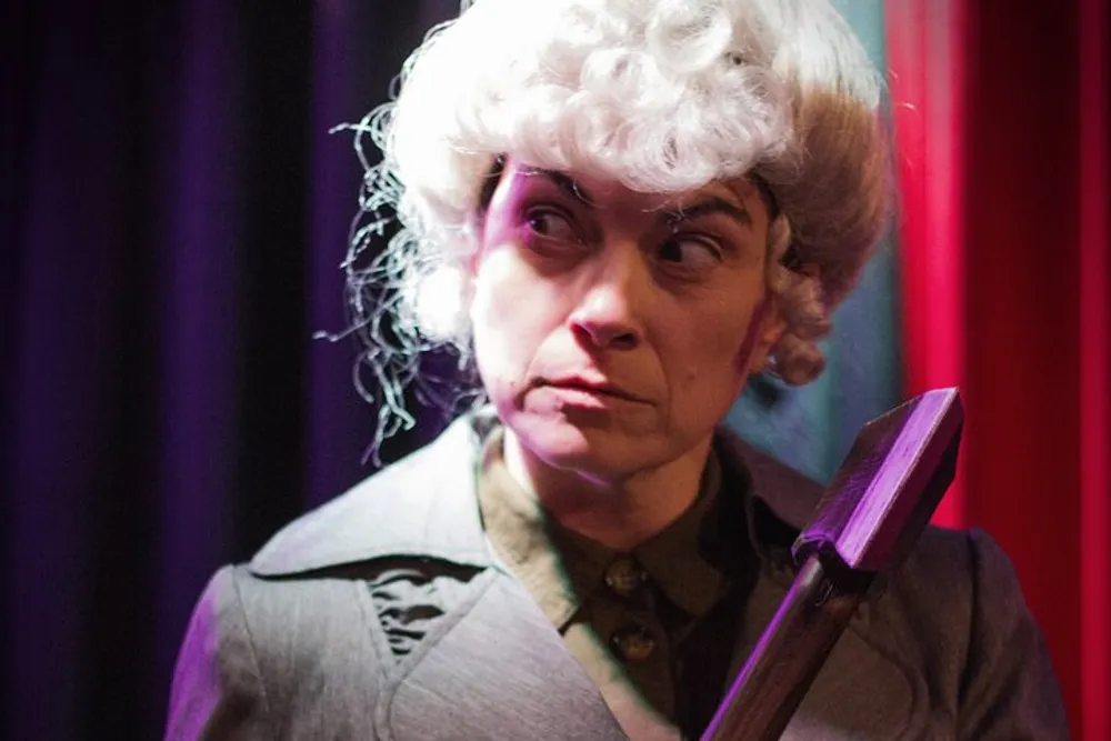 A person is wearing a wig with curls portraying a dramatic expression and holding a prop that resembles a walking cane or a weapon
