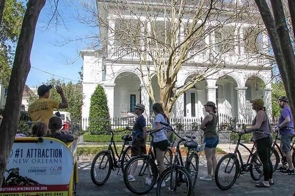 A group of cyclists is stopped to listen to a guide during a bike tour in an urban area with historical houses and lush greenery in the background