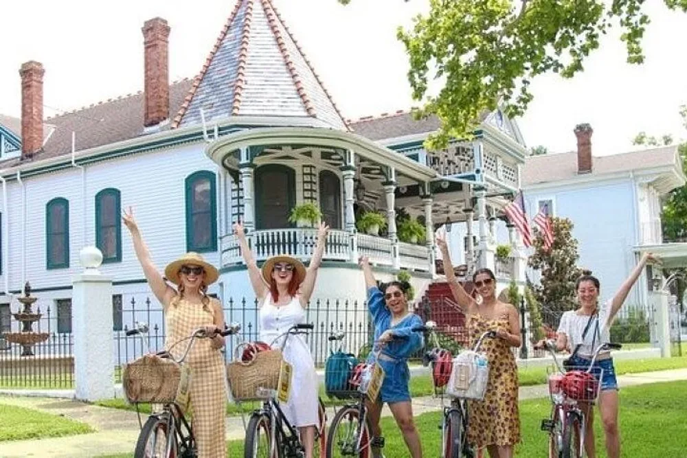 A group of happy individuals are raising their arms cheerfully while standing with bicycles in front of a Victorian-style house with a well-kept lawn