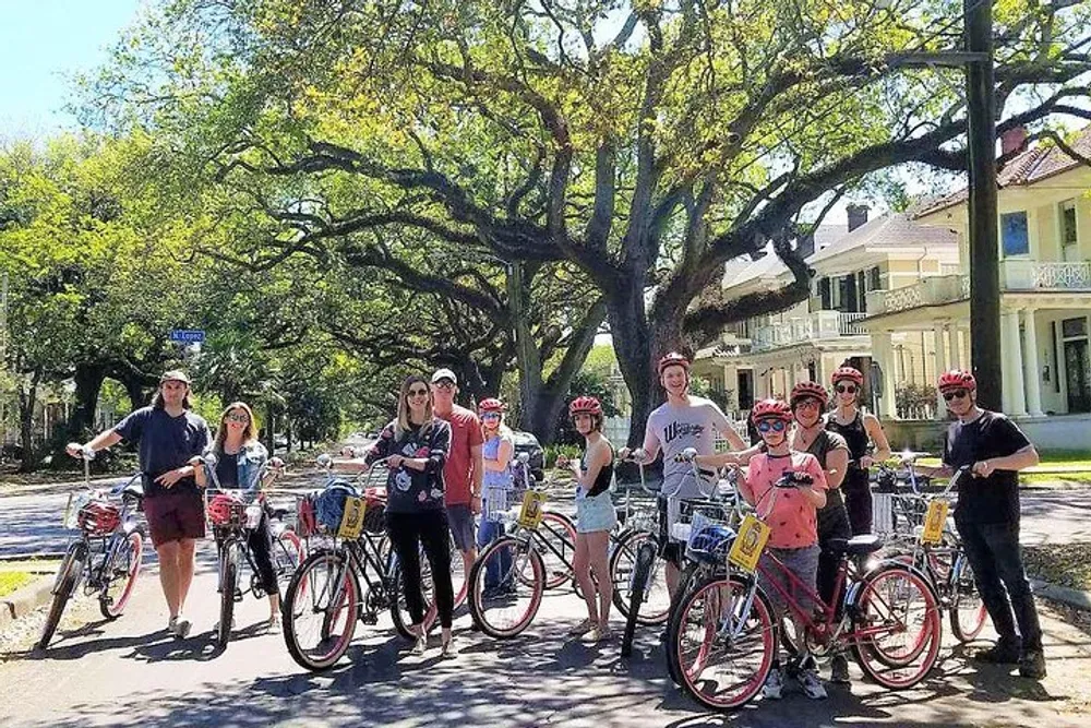 A group of people with bicycles are posing together on a sunny street lined with large leafy trees