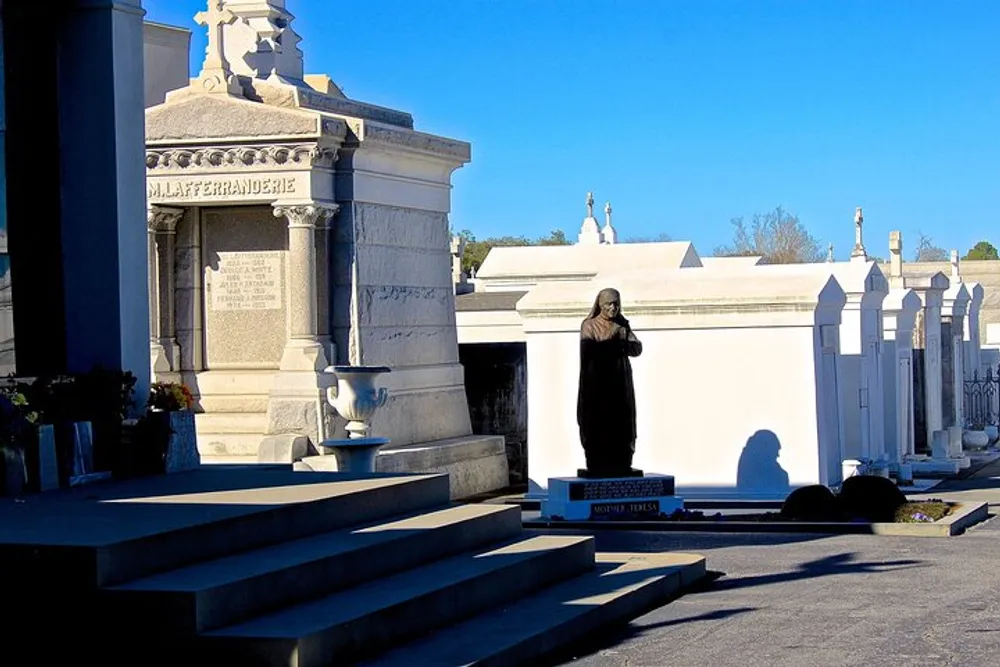 The photograph shows a peaceful and somber scene with a collection of ornate white and gray tombs monuments and a statue within a sunlit cemetery