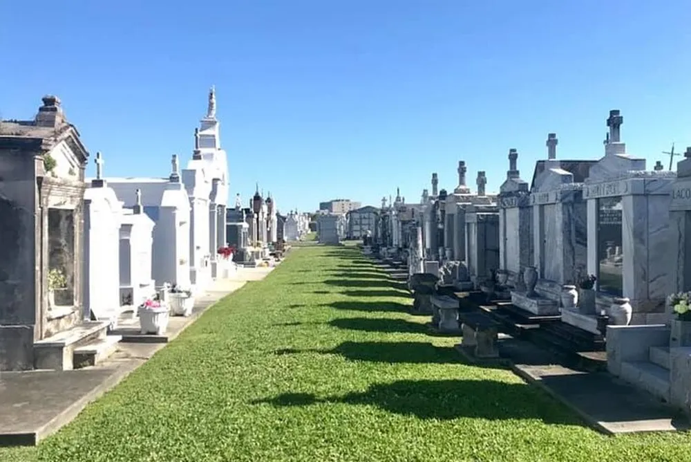 The image shows a sunny view of an expansive cemetery with rows of ornate mausoleums and tombs characterized by a green lawn pathway under a clear blue sky