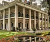 The image shows an elegant southern-style mansion with tall columns a second-floor balcony and surrounded by lush gardens
