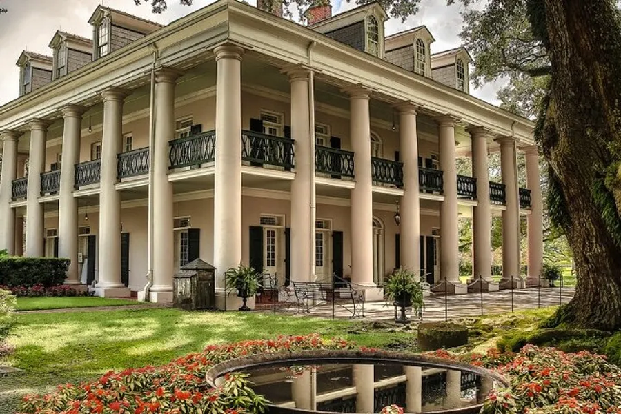 The image shows an elegant southern-style mansion with tall columns, a second-floor balcony, and surrounded by lush gardens.