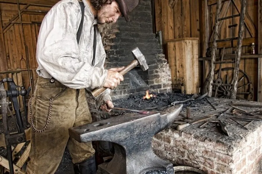 A person dressed in historical clothing is working with a hammer and anvil in a blacksmith's forge.