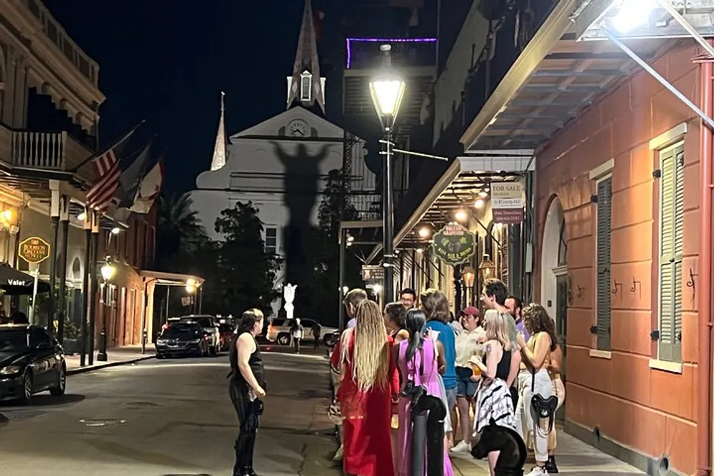 A group of people some in costumes are gathered on a well-lit street at night with historic architecture and a church spire in the background